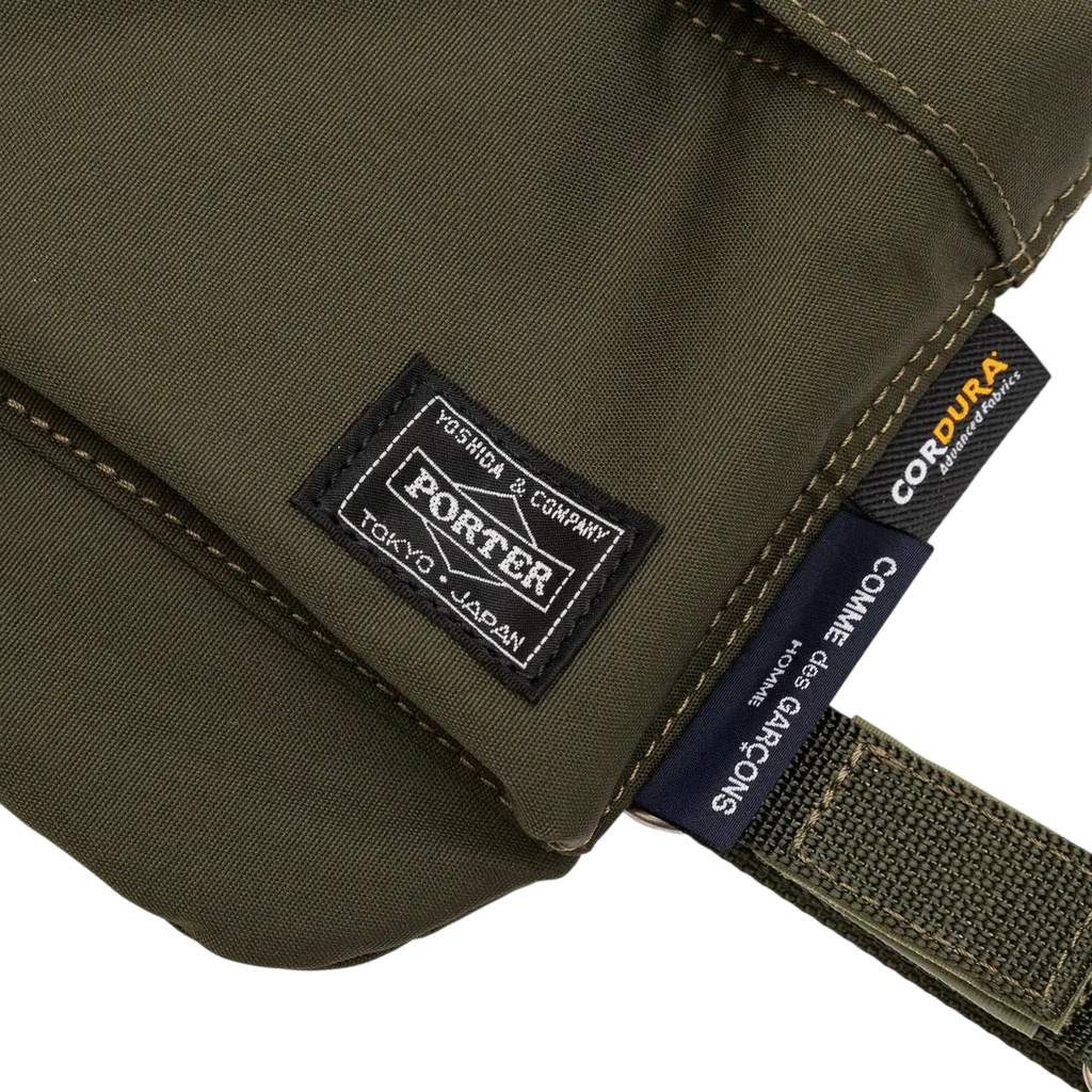 Olive green canvas shoulder bag with black embroidered brand patch and navy blue label from Comme des Garcons x Porter