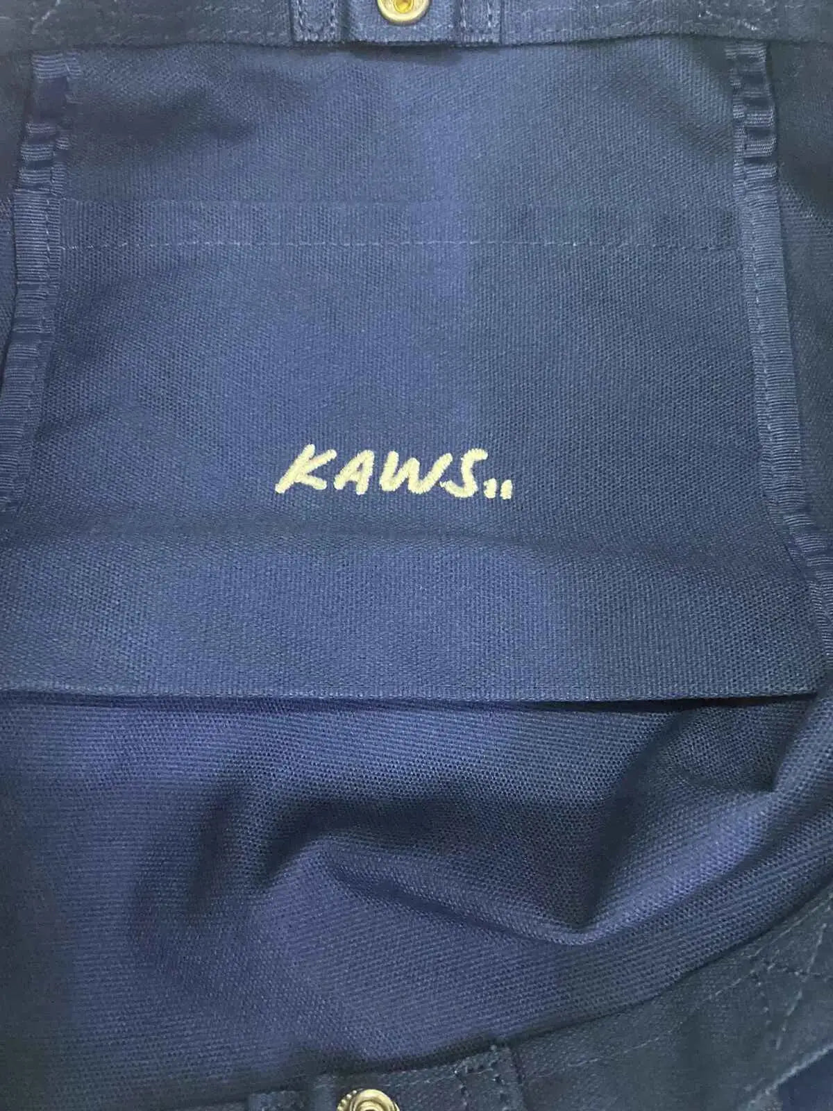 Dark blue canvas backpack with yellow embroidered Kaws logo featured in Kaws x Uniqlo Allover Print Tote Navy collection